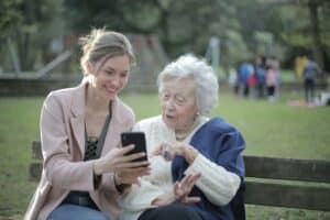a younger woman showing an elderly woman pictures on her phone sitting on a bench. Another reason for investing in assisted living facilities.
